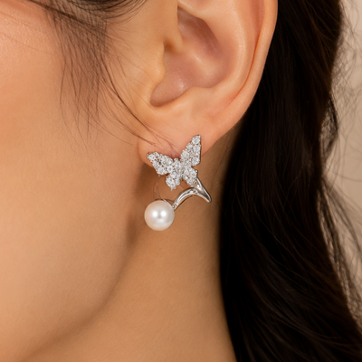 A close-up of a woman's ear wearing a silver butterfly stud earring with a dangling white pearl, against a soft beige background.