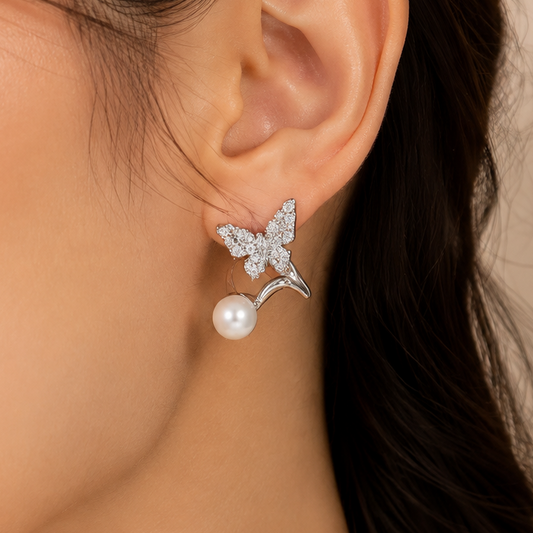A close-up of a woman's ear wearing a silver butterfly stud earring with a dangling white pearl, against a soft beige background.
