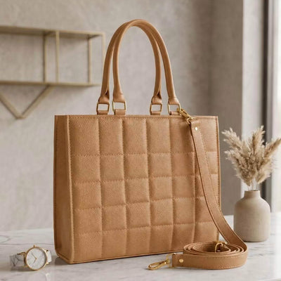 A tan quilted tote bag with double top handles and a matching detachable shoulder strap, styled on a marble countertop with a watch and dried grass in a vase in the background.